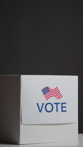 Vertical-Video-Shot-Of-Ballot-Box-With-USA-Flag-In-American-Election-Against-Black-Background